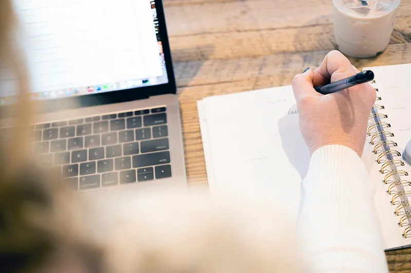 Une personne écrivant dans un carnet à côté d'un ordinateur portable, symbolisant la rédaction stratégique pour le SEO