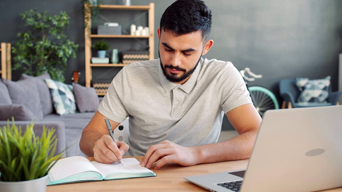 Un homme écrivant des notes dans un carnet à côté de son ordinateur, symbolisant la recherche et la rédaction en copywriting